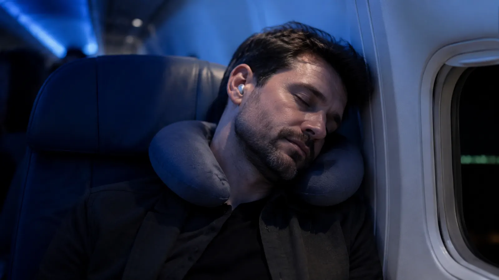 Traveler asleep on their side against an airplane window with a small silicone earplug visible at the outer ear