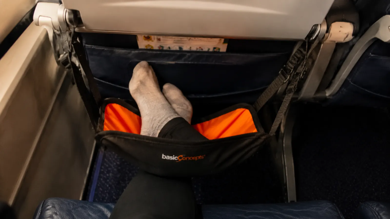Close-up of a BASIC CONCEPTS foot hammock strapped under an airplane tray table with feet resting on it
