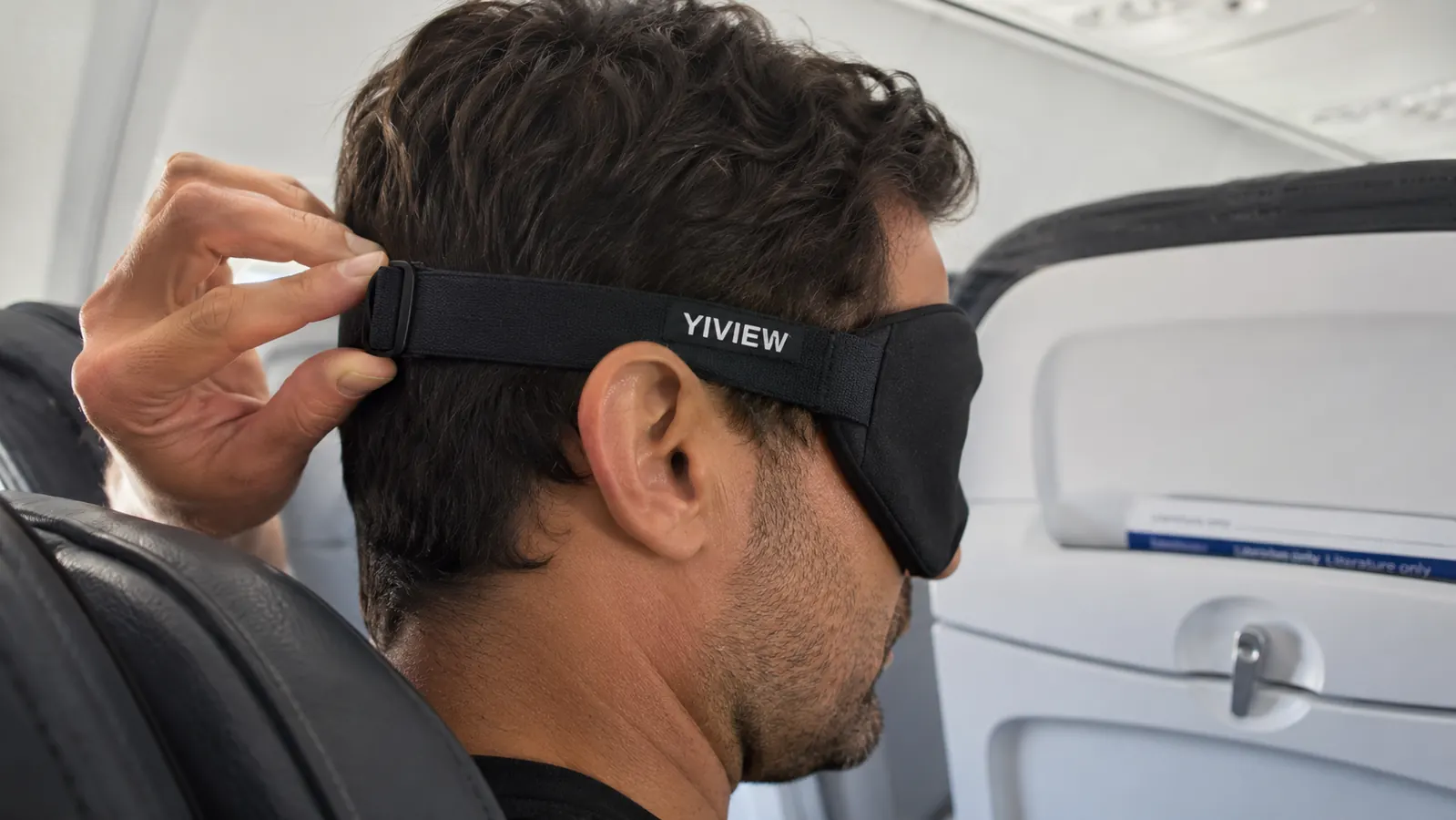 Hand adjusting the strap of a 3D contoured sleep mask before reclining an airplane seat
